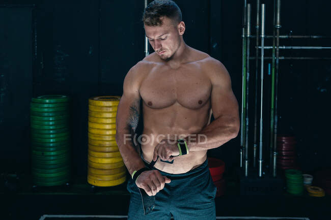 Впевнений спортсмен з м'язистим голим торсом стоїть в тренажерному залі і обгортає зап'ястя бинтами під час підготовки до функціональних тренувань — стокове фото