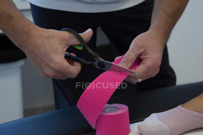 Physiothérapeute masculin en masque stérile coupant la bande élastique de kinésiologie avec des ciseaux à l'hôpital — Photo de stock