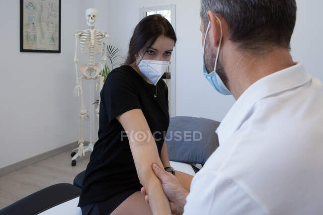 Quiroprático masculino anônimo em máscara respiratória examinando antebraço da mulher durante o processo de fisioterapia no centro médico — Fotografia de Stock