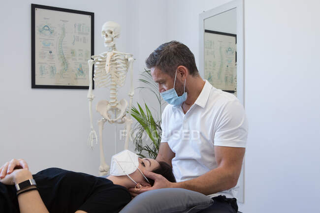 Fisioterapeuta masculino irreconhecível em máscara facial verificando o pescoço da mulher perto do esqueleto humano no centro médico — Fotografia de Stock