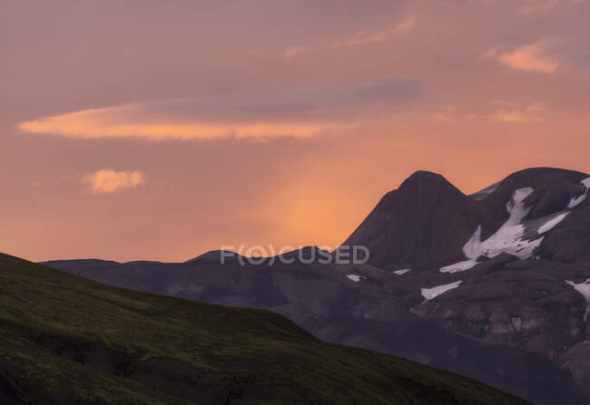 Impresionante vista de majestuosas montañas con nieve en las laderas cerca del valle de colinas cubiertas de hierba bajo el pintoresco cielo rosado de la noche en Islandia - foto de stock