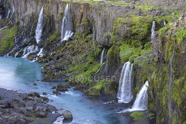 Espectacular vista de las rápidas cascadas que fluyen desde el áspero acantilado rocoso cubierto de exuberante vegetación hacia el tranquilo embalse azul en naturaleza pacífica - foto de stock