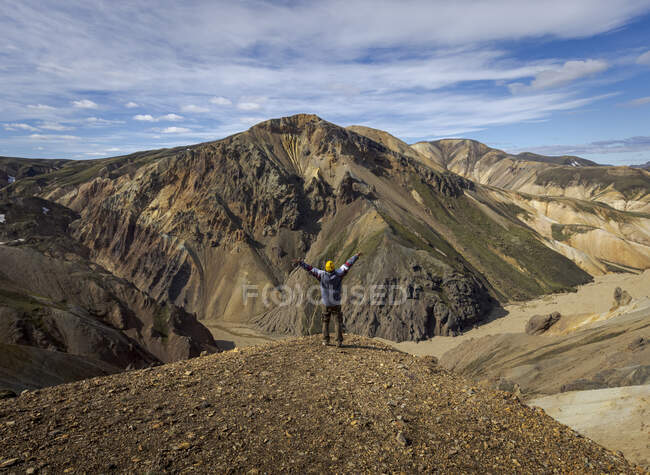 Caminante masculino con traje casual con los brazos extendidos en la emoción de pie en la colina seca contra el pintoresco terreno montañoso en el día soleado - foto de stock