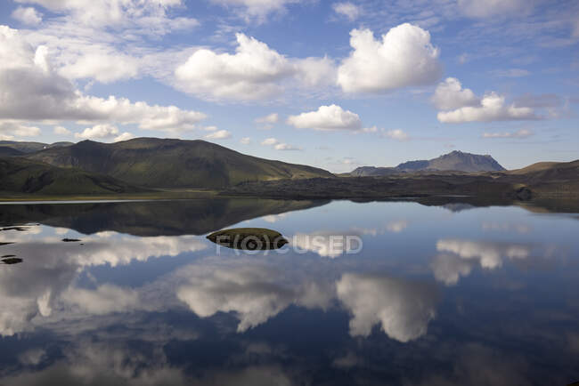 Paysage à couper le souffle d'un lac encore tranquille reflétant un ciel bleu clair et entouré de collines verdoyantes rocheuses dans des hauts plateaux paisibles en Islande — Photo de stock