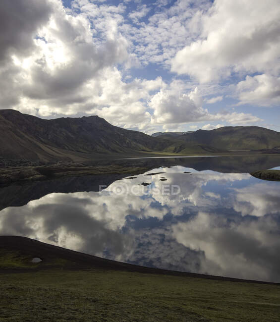 Impresionante paisaje de lago todavía tranquilo que refleja el cielo azul claro y rodeado de colinas verdes rocosas en tierras altas pacíficas en Islandia - foto de stock