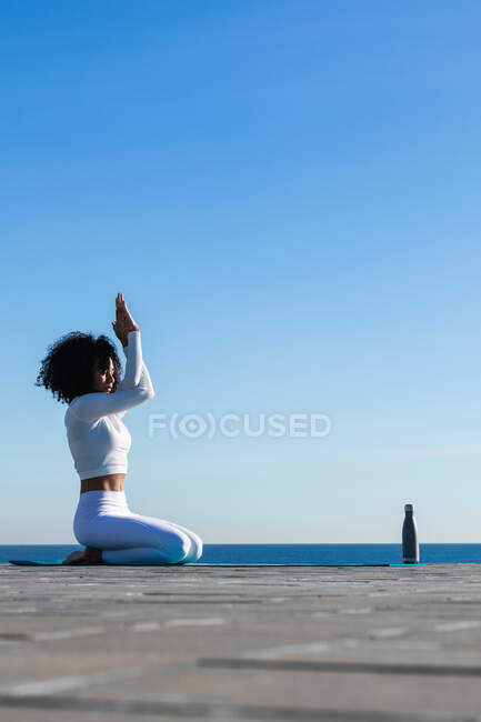 Side view of fit female with curly hair dressed in sportswear doing Eagle Pose while sitting on knees on sports mat with reusable water bottle on the seaside — Stock Photo
