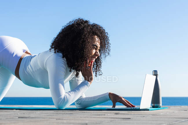 Side view full body of positive ethnic female with legs up watching netbook during yoga session on waterfront — Stock Photo