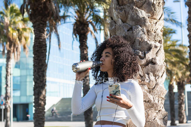Young fit Brazilian female athlete with curly hair drinking water from zero waste bottle and listening to playlist on smartphone after yoga training — Stock Photo