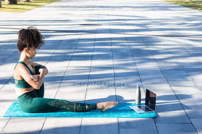 Vista lateral de cuerpo completo de mujer brasileña haciendo ejercicio de estiramiento mientras ve tutorial de yoga en netbook en el parque - foto de stock