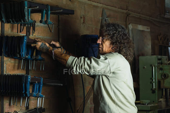 Seitenansicht eines reifen männlichen Tischlers, der in einer schäbigen Holzwerkstatt eine Schreinerklammer aus dem Regal holt — Stockfoto