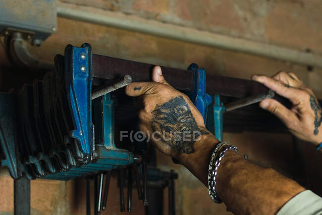 Seitenansicht eines bis zur Unkenntlichkeit geschnittenen männlichen Tischlers, der in schäbiger Holzwerkstatt eine Schreinerklammer aus dem Regal holt — Stockfoto