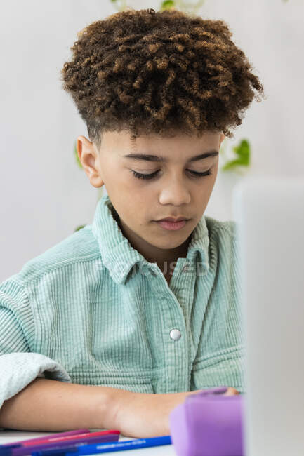 Ретельний афроамериканський школяр у повсякденній сорочці, який сидить за столом і записує в підручнику під час роботи над домашнім завданням. — стокове фото