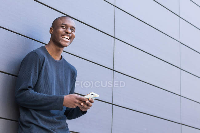 Side view of happy African American male scrolling modern mobile phone while standing against gray building — Stock Photo