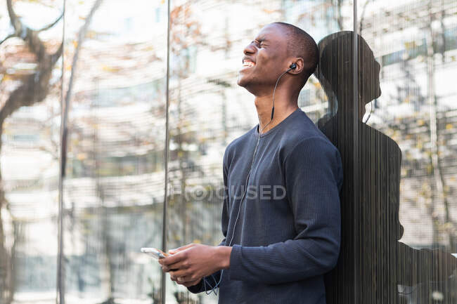 Joyful African American man enjoying music in earphones while using cellphone standing near glass wall — Stock Photo