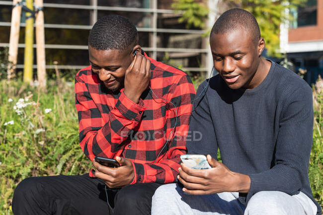 Cheerful African American male friends listening to music together on mobile phone while sitting in park together — Stock Photo