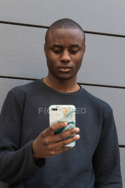 Attento afroamericano maschio scorrimento moderno telefono cellulare mentre in piedi contro edificio grigio — Foto stock