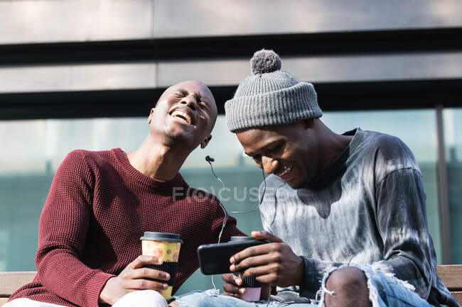 Young content best African American friends with coffee to go and earphones watching cellphone in city — Stock Photo