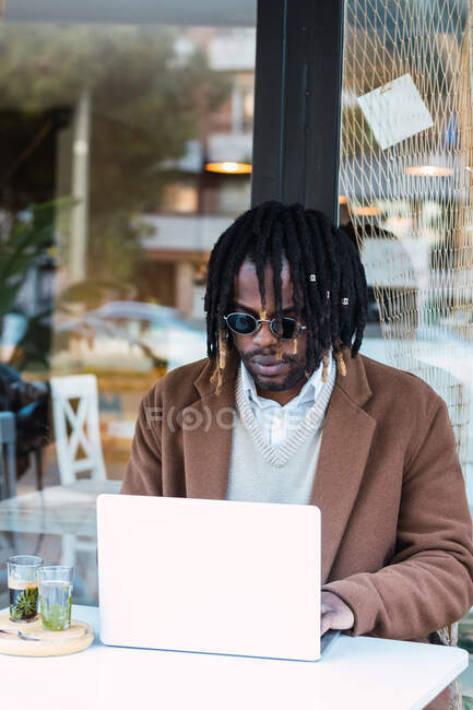 Grave focalizzato hipster maschio nero in occhiali da sole con dreadlocks seduto a tavola con computer portatile mentre si lavora su un progetto remoto in ristorante all'aperto — Foto stock