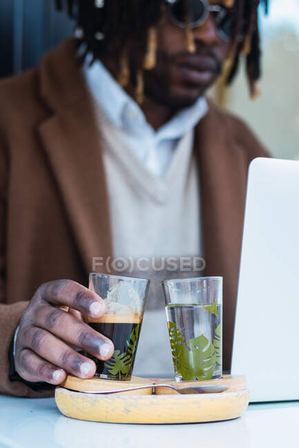 Unemotional blurred African American male sitting at table with glass of water and espresso in street coffee shop working on laptop — Stock Photo