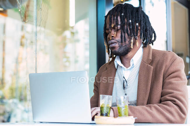 Серьезный ориентированный хипстер черный мужчина с афро в дредах сидит за столом с ноутбуком во время работы над удаленным проектом в открытом ресторане — стоковое фото