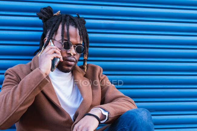 Grave elegante maschio afro-americano con trecce afro che indossano abiti alla moda e occhiali da sole che hanno conversazione telefonica mentre seduto contro la parete di metallo blu — Foto stock