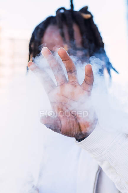 Hombre afroamericano irreconocible con rastas cubriendo la cara con la mano mientras está parado en la calle en la nube de humo y fumando cigarrillo electrónico - foto de stock