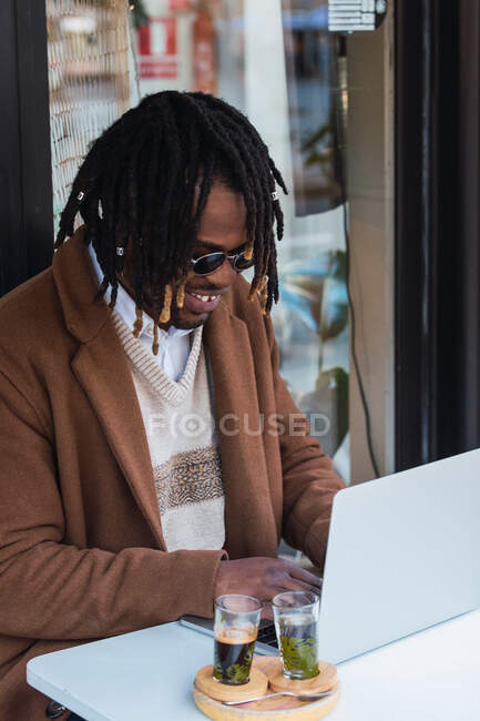 Vista laterale di allegro hipster focalizzato maschio nero con Afro in dreadlocks seduto a tavola con computer portatile mentre si lavora su un progetto remoto nel ristorante all'aperto — Foto stock