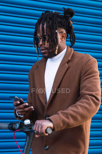Branché hipster afro-américain gars en manteau élégant et avec dreadlocks navigation téléphone mobile tout en chevauchant scooter électrique contre mur bleu — Photo de stock