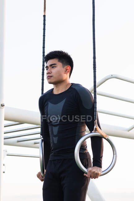 Low angle of young Asian sportsman in black activewear hanging on gymnastic rings while performing exercises on sports ground — Stock Photo