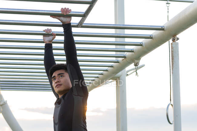 Strong determined young Asian man in sportswear doing exercises on monkey bars during outdoor workout on sports ground at sunset — Stock Photo