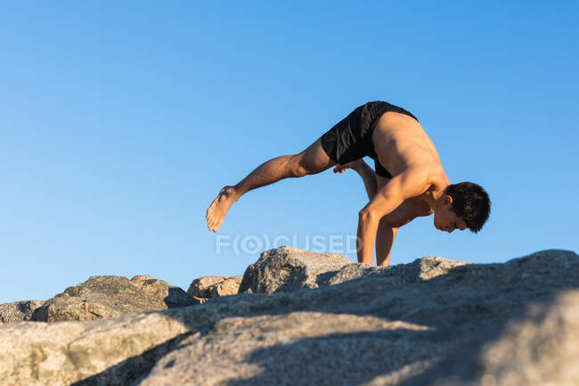 Vista laterale del corpo pieno di flessibile focalizzato giovane maschio etnico con torso nudo praticando avanzato Flying Pigeon asana durante la sessione di yoga su terreno roccioso contro il cielo blu — Foto stock