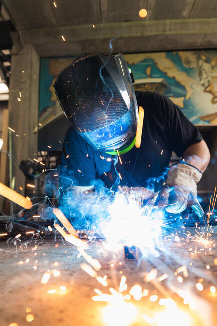 Unrecognizable male master in protective helmet welding metal piece at workbench in workshop — Stock Photo