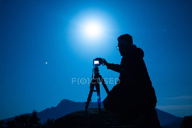 Side view of unrecognizable male traveler silhouette with photo camera on tripod against ridge under blue sky with shiny sun at night — Stock Photo