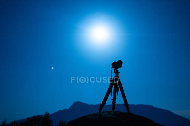 Профессиональная фотокамера с ремнем на штативе против горного силуэта под красочным небом с солнцем в сумерках — стоковое фото