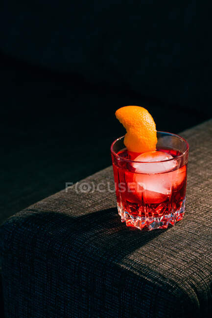 Copa de refrescante cóctel Negroni con sabor amargo y hielo adornado con cáscara de naranja y servido en el brazo del sofá en habitación oscura - foto de stock