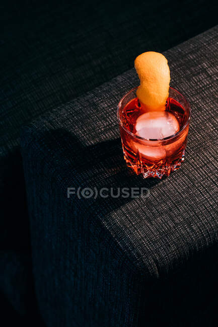 Glass of refreshing Negroni cocktail with bitter flavor and ice garnished with orange peel and served on couch arm in dark room — Stock Photo