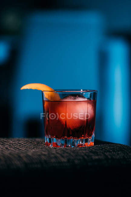 Стакан освежающего коктейля Негрони с горьким вкусом и льдом, украшенный апельсиновой кожурой и подаваемый на диване в темной комнате — стоковое фото