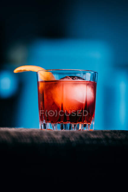 Bicchiere di rinfrescante cocktail Negroni dal sapore amaro e ghiaccio guarnito con buccia d'arancia e servito sul braccio del divano in camera oscura — Foto stock