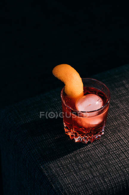 Bicchiere di rinfrescante cocktail Negroni dal sapore amaro e ghiaccio guarnito con buccia d'arancia e servito sul braccio del divano in camera oscura — Foto stock