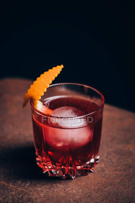 Стакан освежающего коктейля Негрони с горьким вкусом и льдом, украшенный апельсиновой кожурой и подаваемый на диване в темной комнате — стоковое фото