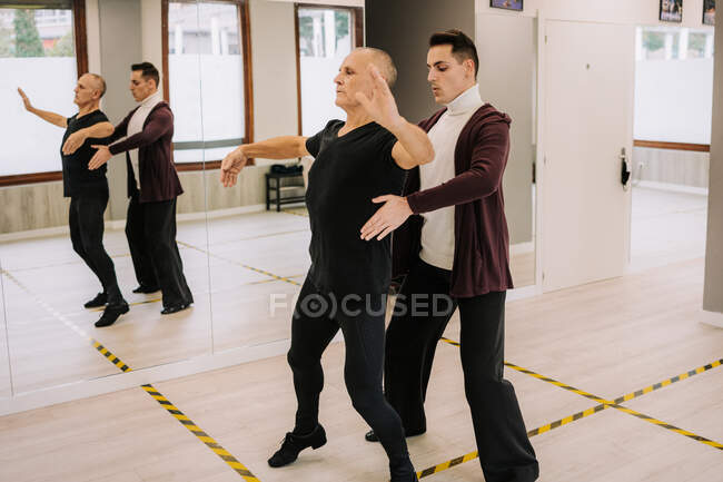 Боковой вид мужчины-инструктора, помогающего зрелой танцовщице с движением во время бальных танцев в просторном зале — стоковое фото