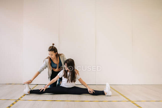Entrenador femenino sonriente en ropa deportiva que ayuda a una chica alegre con piernas estiradas mientras se calienta antes del entrenamiento físico - foto de stock