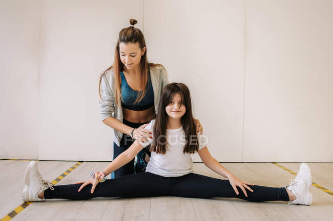 Treinador feminino sorridente em sportswear ajudando menina alegre com pernas esticando enquanto se aquece antes do treino de fitness — Fotografia de Stock