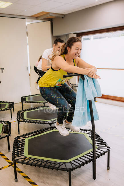 Compañía de atletas que saltan activamente sobre el suelo en el trampolín mientras hacen ejercicios durante el entrenamiento activo de la aptitud en el gimnasio moderno brillante - foto de stock