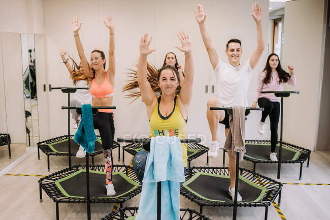Company of sportspeople jumping on trampolines with raised arms during active fitness training in gym — Stock Photo