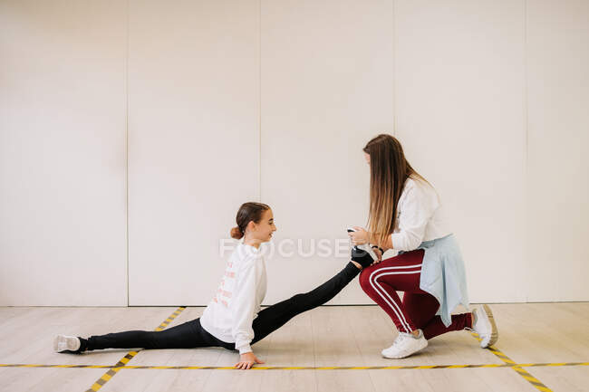 Вид сбоку гибкой девочки, сидящей в расколотых и растянутых ногах с помощью тренера по танцам перед уроком в просторном зале — стоковое фото