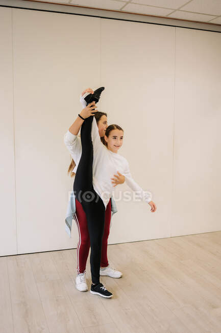 Flexible girl standing in split and stretching legs with help of dance trainer before lesson in spacious hall — Stock Photo