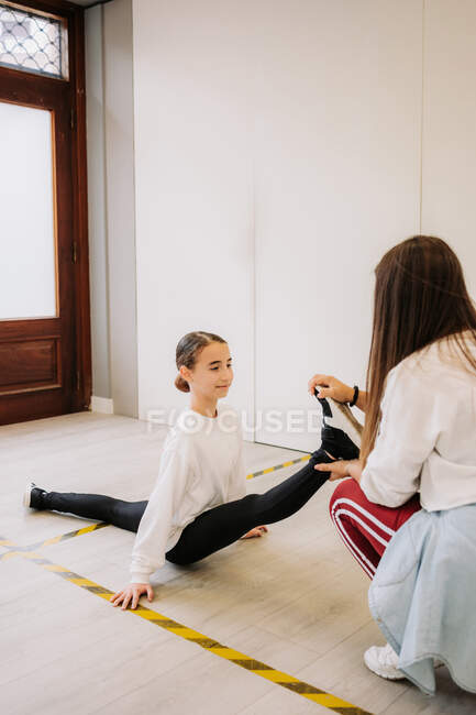 Vista lateral de una chica flexible sentada en piernas abiertas y estiradas con la ayuda de un entrenador de baile antes de la lección en un amplio salón - foto de stock