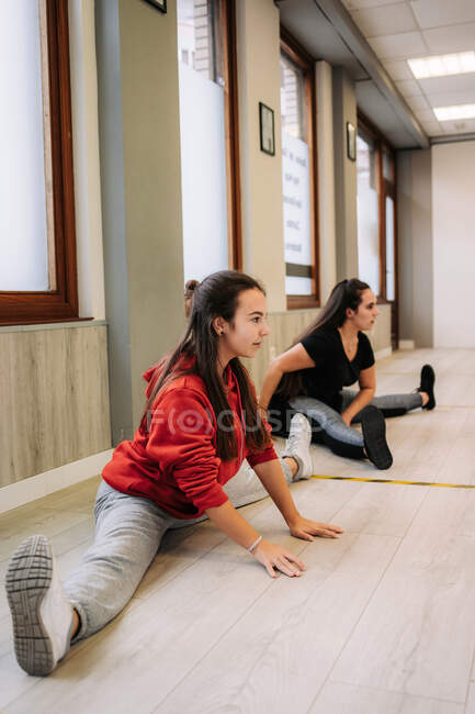 Entrenador femenino sonriente en ropa deportiva que ayuda a una chica alegre con piernas estiradas mientras se calienta antes del entrenamiento físico - foto de stock