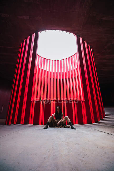 Ethnische Theaterkünstlerin sitzt breitbeinig auf dem Boden in der Nähe einer hellen Rippenwand auf der Bühne und blickt in die Kamera — Stockfoto